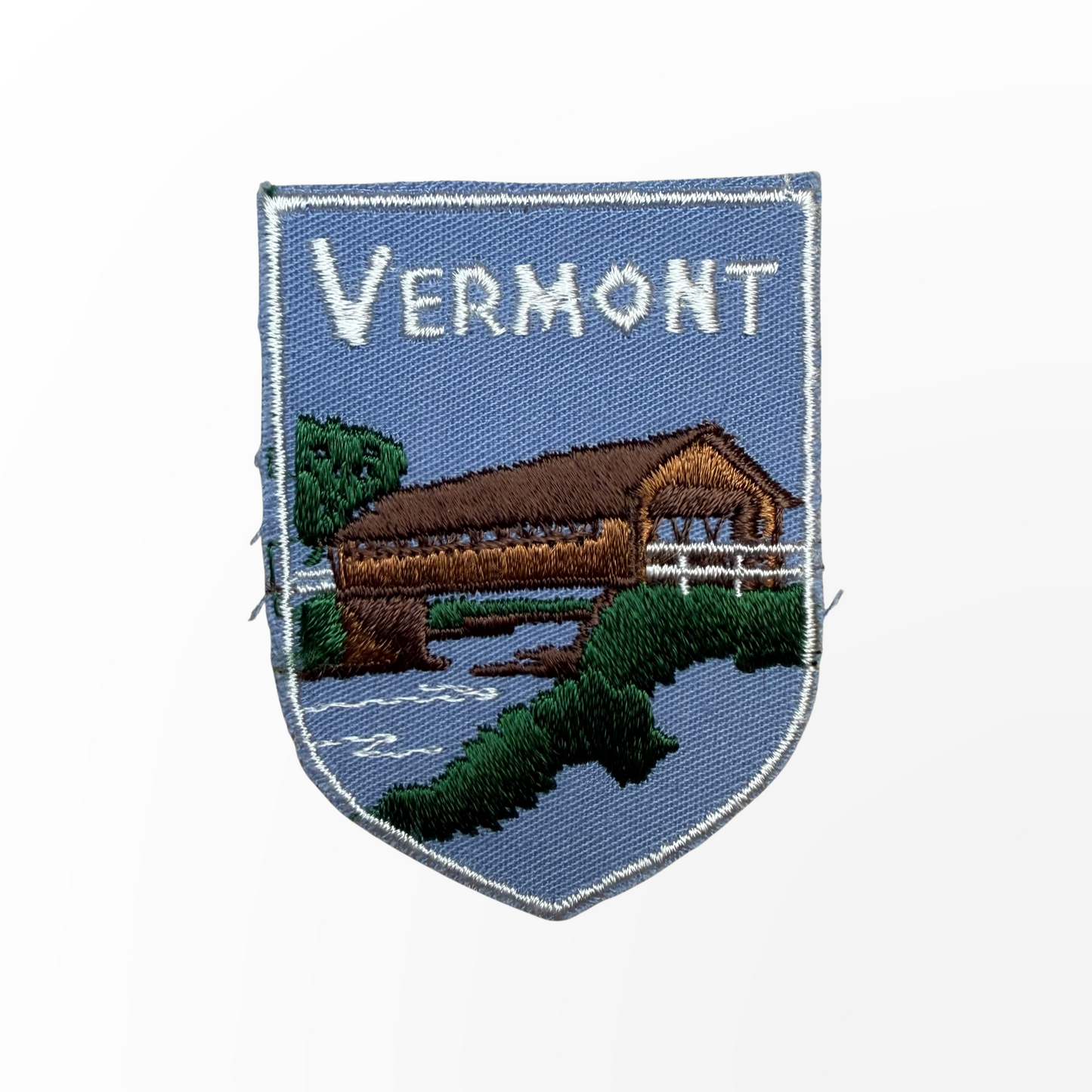 An embroidered shield-shaped patch featuring a Vermont landscape with a covered bridge, trees, water, and the word 'VERMONT' in white text on a blue background.