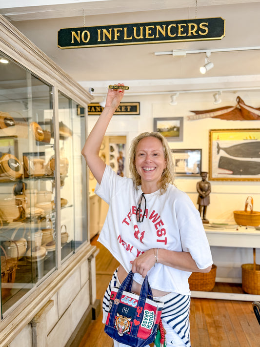 A woman smiling in a museum or shop interior, holding up a small gold-colored object in one hand while wearing a white graphic T-shirt, striped bottoms, and carrying a blue patterned tote bag. A hanging sign above reads 'NO INFLUENCERS.