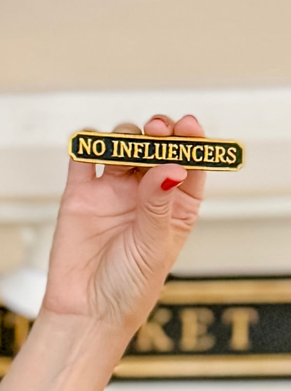 A hand with red nail polish holds a small rectangular enamel pin that reads 'NO INFLUENCERS' in gold lettering on a black background, with a softly blurred indoor setting behind it.