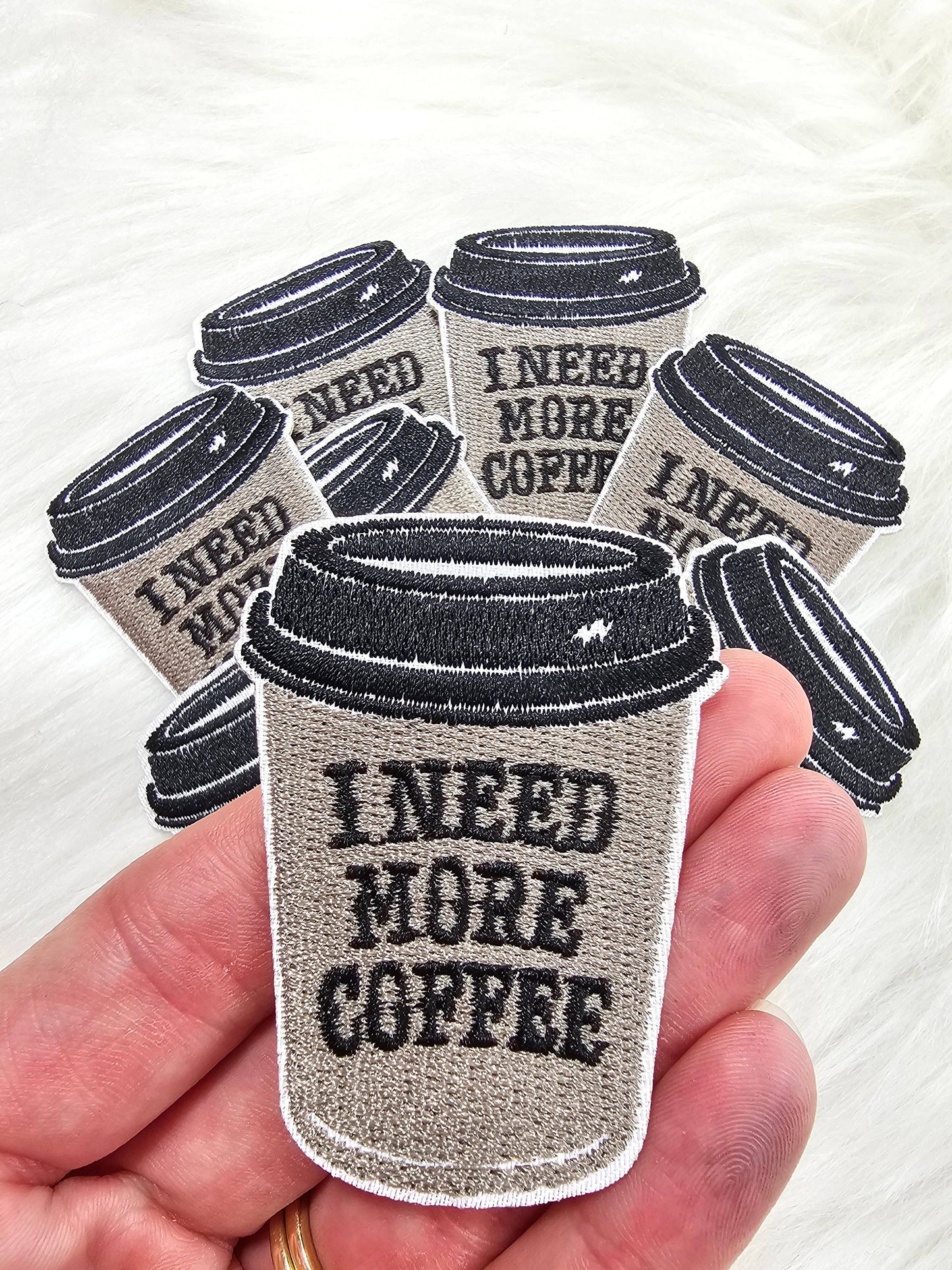 Patch of a coffee cup with 'I Need More Coffee' text held in a hand.