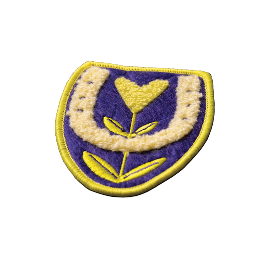 Embroidered patch with a yellow heart and leaves on a purple background, placed on a textured fabric surface.