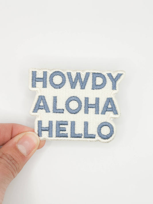 Embroidered patch with 'HOWDY ALOHA HELLO' text held by a hand on a white background