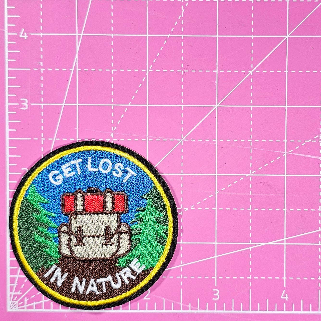 Embroidered patch with 'Get Lost in Nature' text on a pink cutting mat.