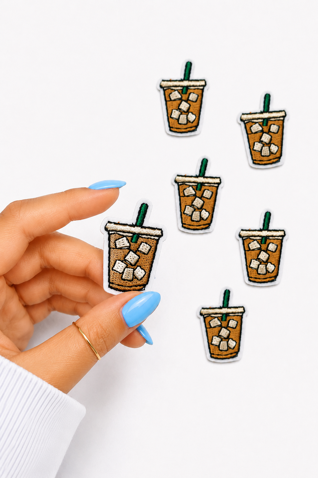 A hand with light blue manicured nails holds an embroidered patch of an iced coffee cup, with five identical patches scattered on a plain white background.