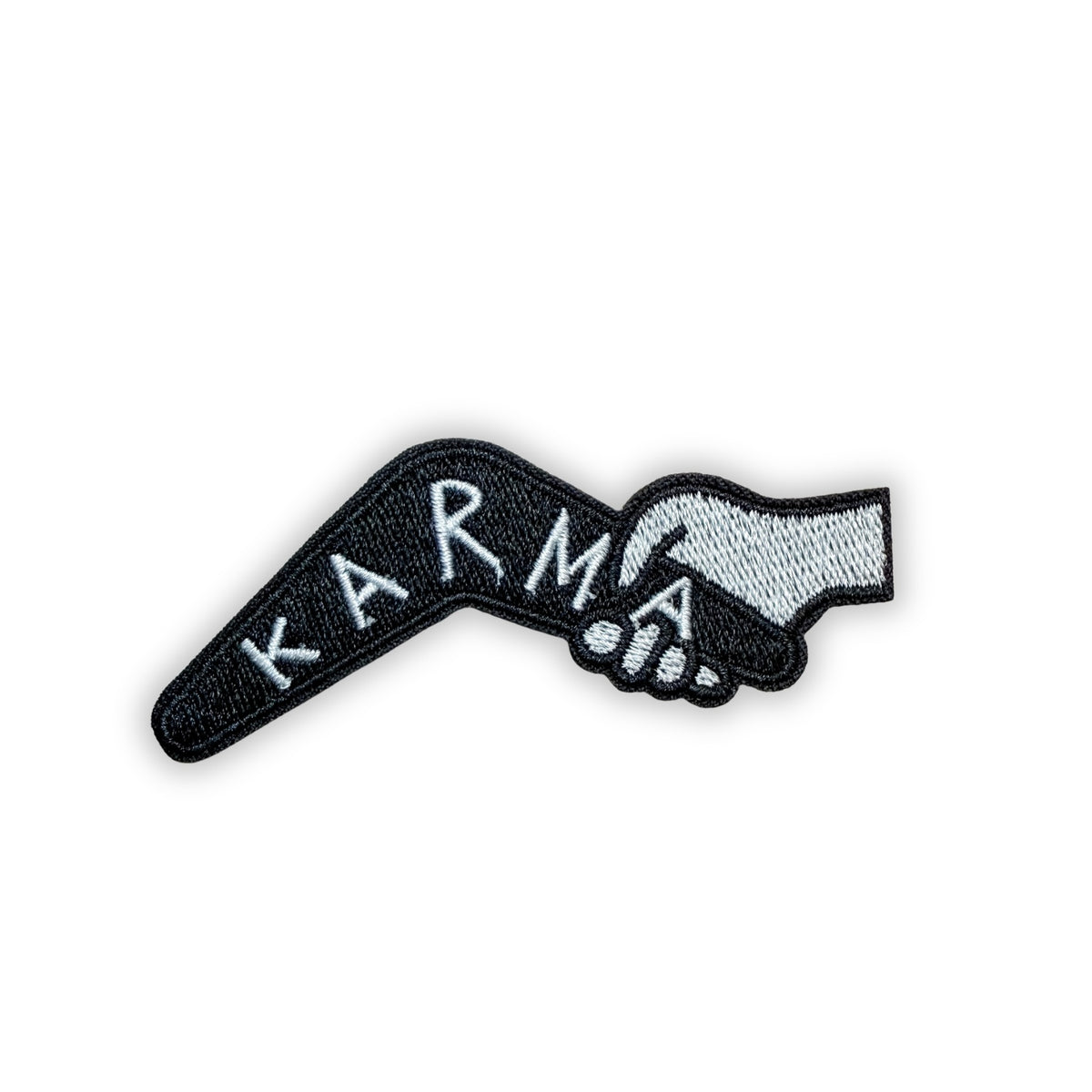 Black embroidered boomerang patch with "KARMA" in white hand-lettered capitals, held at one end by a gray hand, on a white background.