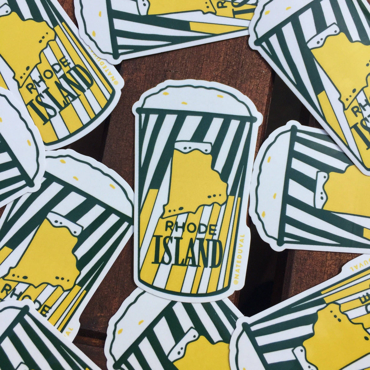 A close-up of multiple die-cut stickers featuring a beer cup illustration with the text 'RHODE ISLAND' and a yellow map outline, arranged on a brown wooden surface.