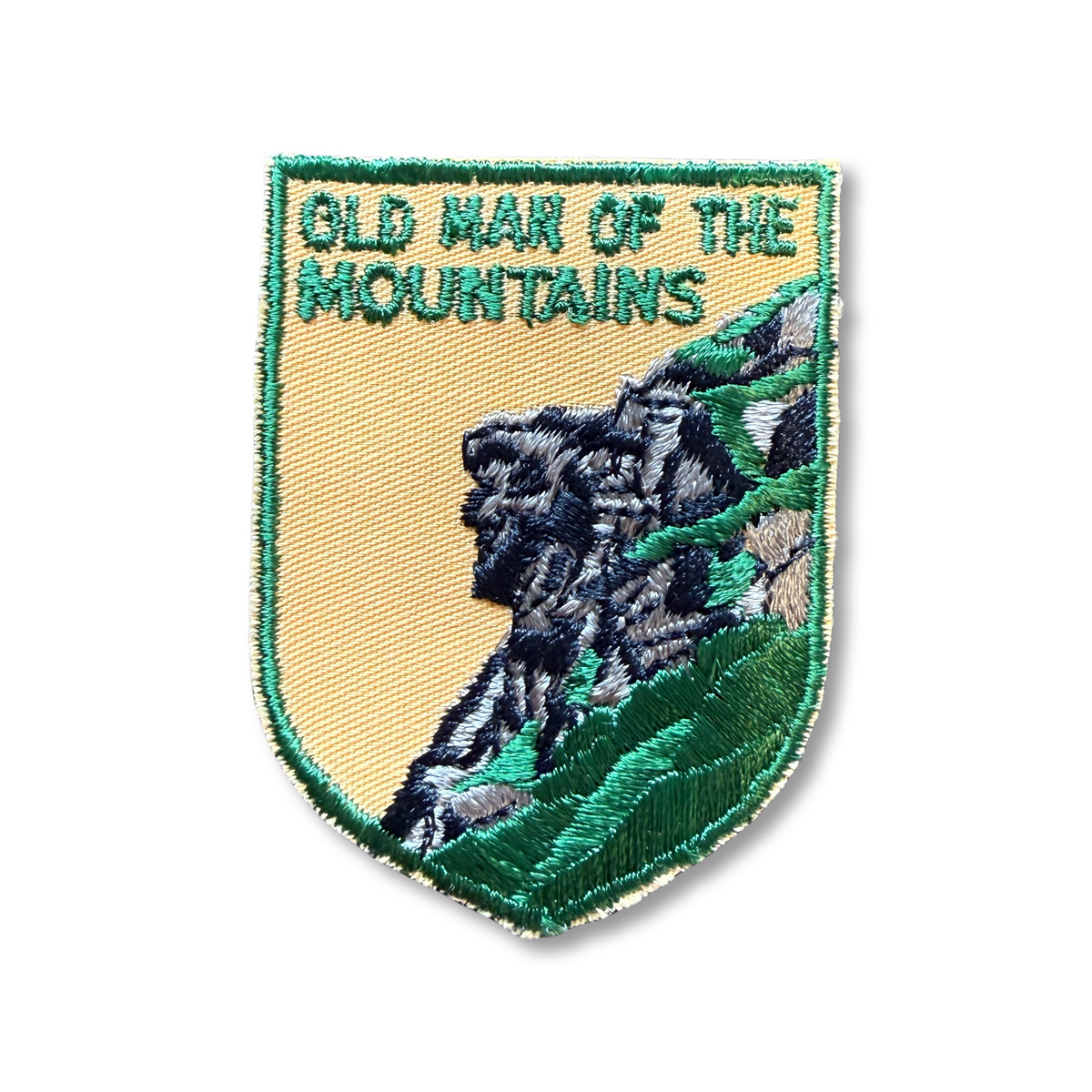 Vintage shield-shaped embroidered patch reading "Old Man of the Mountains" in green letters on goldenrod twill. It shows the Old Man rock profile stitched in black and gray against a green hillside, with a green merrowed border.