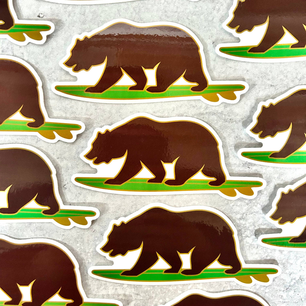 A repeating pattern of die-cut stickers featuring a brown bear walking on a green grassy patch, shown on a light gray textured background.