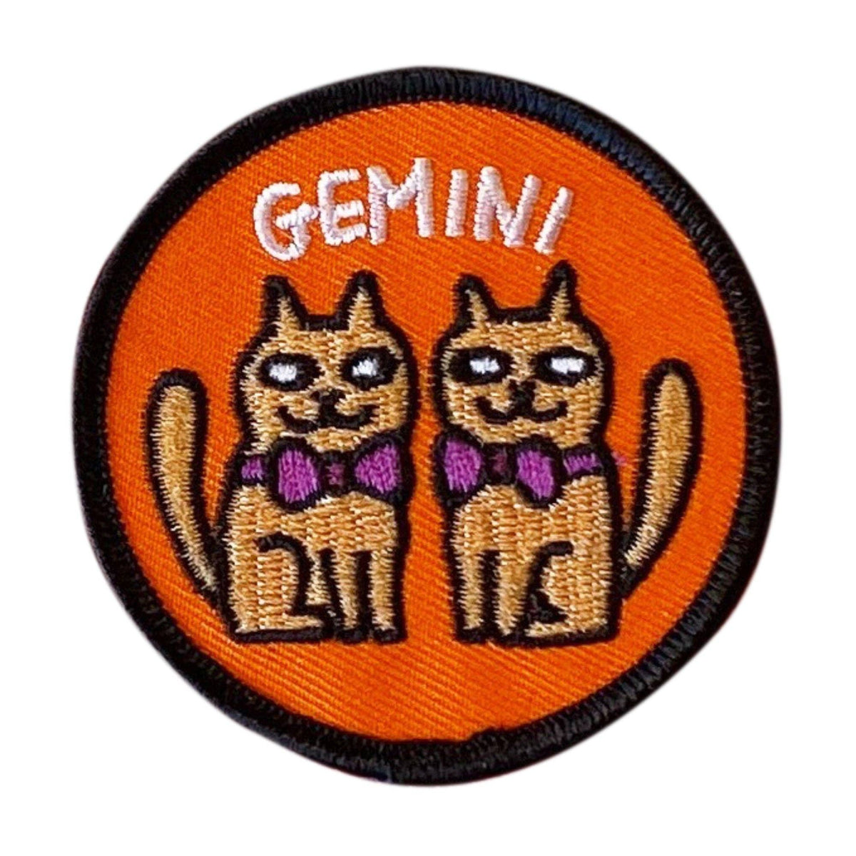 A circular embroidered patch featuring two cartoon cats wearing purple bows, with the word 'GEMINI' in white above them, on an orange background with a black stitched border.