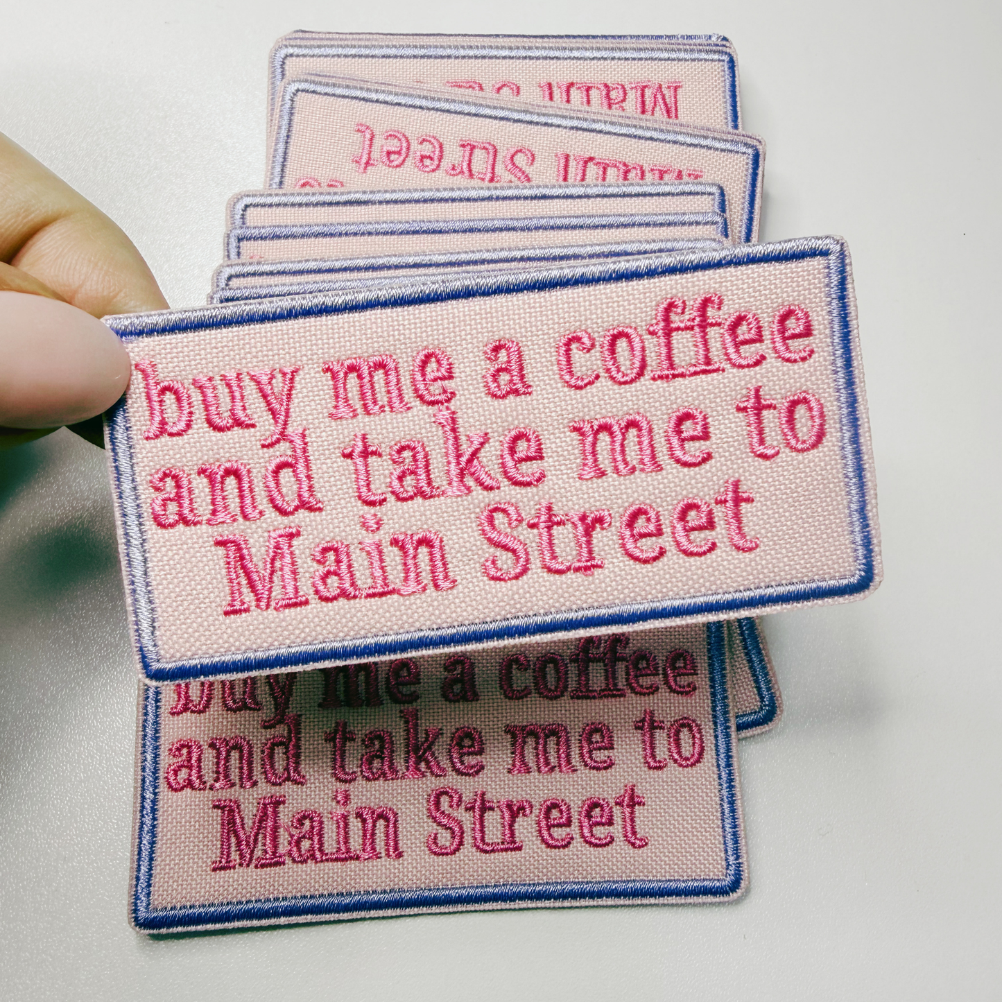 Embroidered patches with text 'buy me a coffee and take me to Main Street' held by a hand.