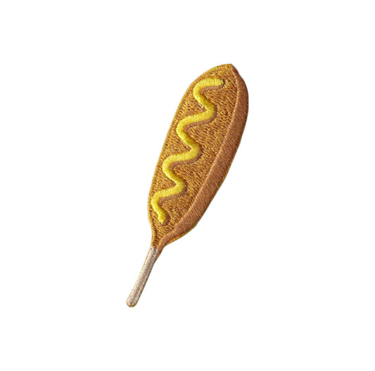An embroidered patch shaped like a hot dog, featuring a golden-brown textured body with a zigzag yellow mustard line and a light beige stem, isolated on a white background.