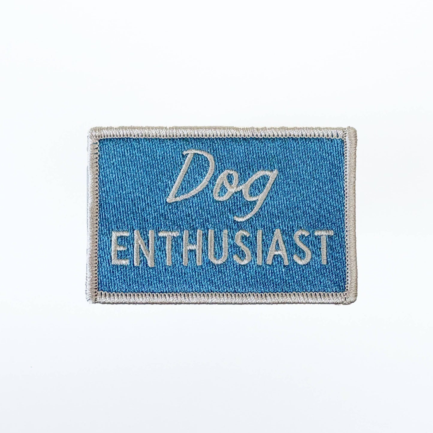 A rectangular embroidered patch with a blue background and a gray border, featuring the words 'Dog ENTHUSIAST' in white lettering, shown on a plain white background.