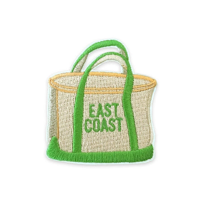 A woven straw tote bag with bright green handles, trim, and fringe, featuring the words 'EAST COAST' embroidered in green on the front, isolated on a white background.