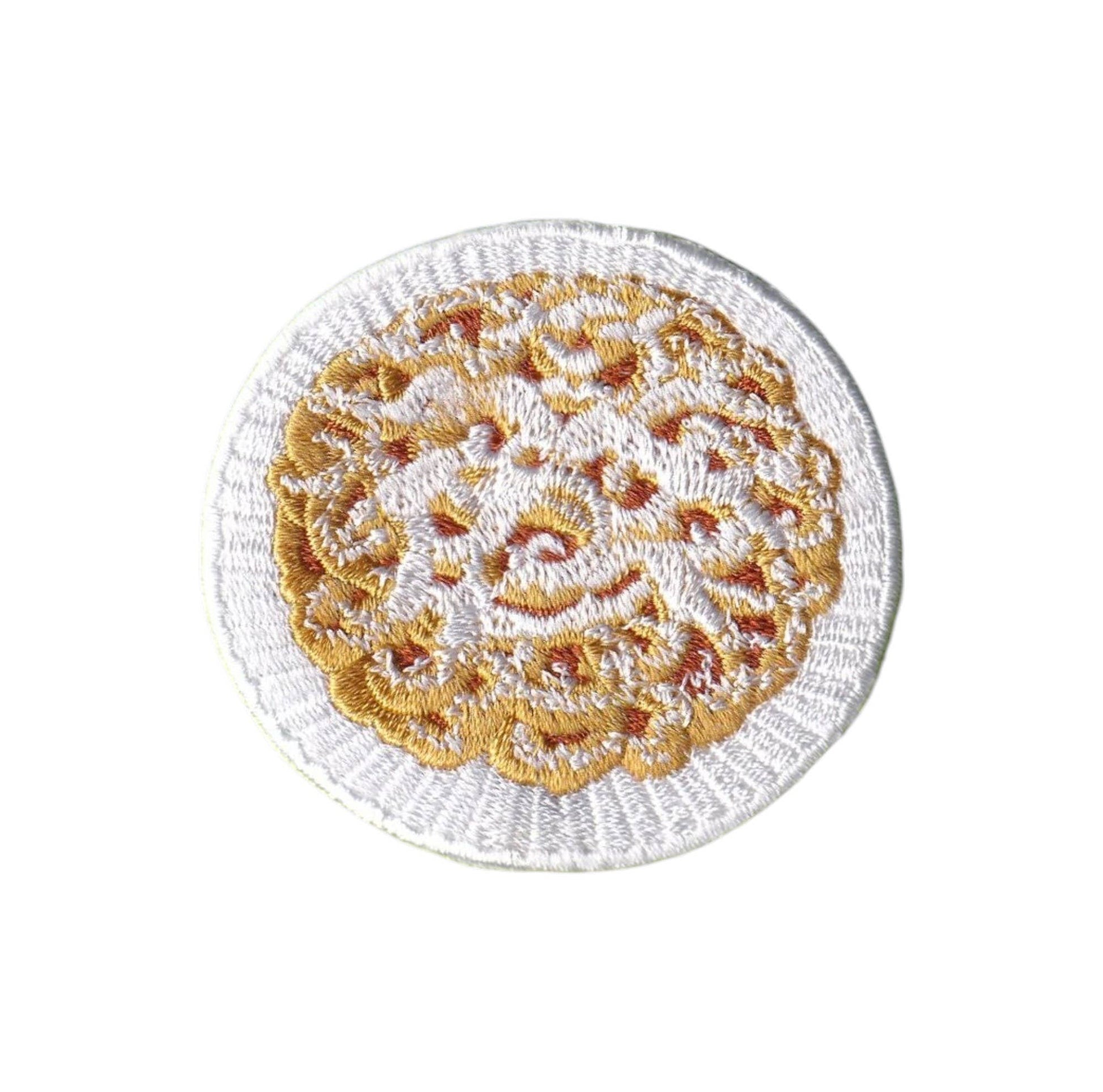 A round embroidered patch featuring a white crochet border and a central gold and orange floral or lace-like pattern on a white background.