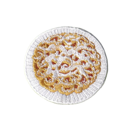 A round embroidered patch featuring a white crochet border and a central gold and orange floral or lace-like pattern on a white background.