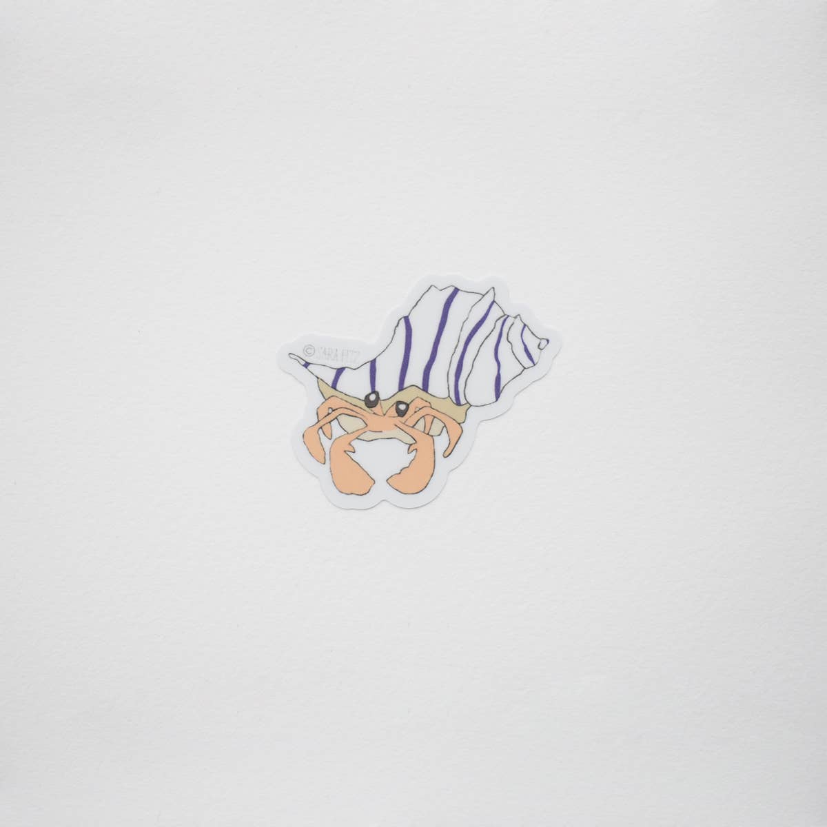 A small die-cut sticker featuring a cartoon hermit crab inside a spiral shell with purple and white stripes, centered on a plain light gray background.