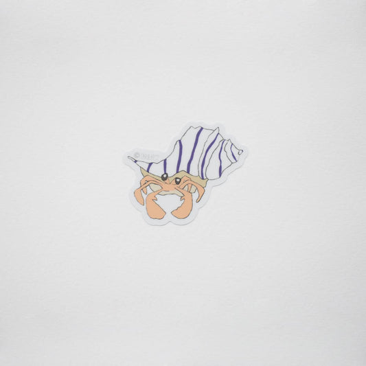 A small die-cut sticker featuring a cartoon hermit crab inside a spiral shell with purple and white stripes, centered on a plain light gray background.