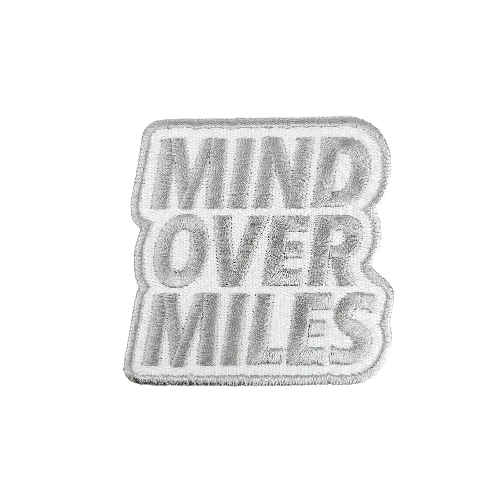 A rectangular embroidered patch with the words 'MIND OVER MILES' in raised gray lettering on a white background, finished with a gray border.