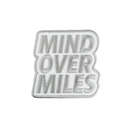 A rectangular embroidered patch with the words 'MIND OVER MILES' in raised gray lettering on a white background, finished with a gray border.