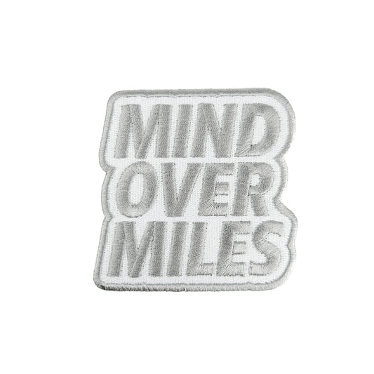 A rectangular embroidered patch with the words 'MIND OVER MILES' in raised gray lettering on a white background, finished with a gray border.