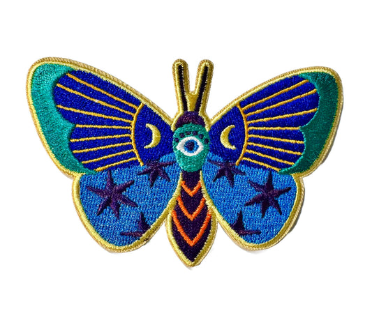 A stylized embroidered moth patch with blue wings, teal edges, gold outlines, and a central eye motif on a plain white background.