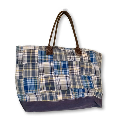 A patchwork plaid tote bag featuring blue, navy, beige, and white fabric squares, with dark brown leather handles and a solid navy bottom panel.