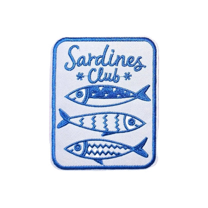A rectangular embroidered patch with rounded corners featuring the words 'Sardines Club' and three stylized blue fish on a white background, placed on a white faux-fur surface.