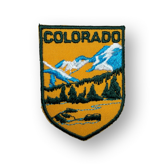 An embroidered shield-shaped Colorado state patch with a yellow-orange field, blue mountains, white snow, dark green trees, and the word "COLORADO" at the top, shown on a white background with a soft shadow.