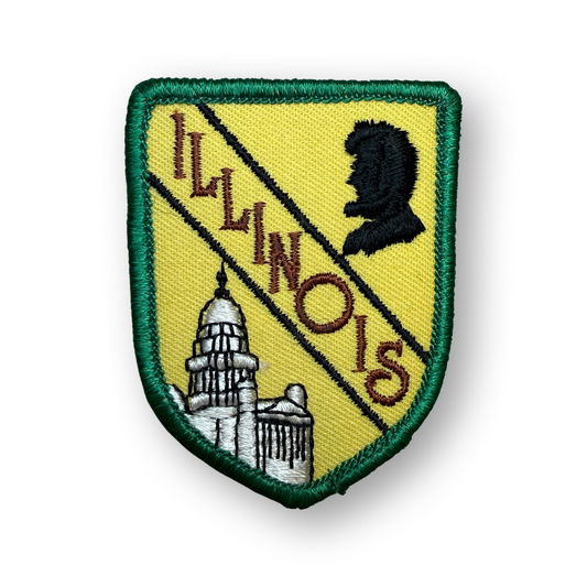 An embroidered Illinois state shield patch with a green border, yellow field, diagonal black stripe, brown 'ILLINOIS' text, a black silhouette profile, and a white-and-gray capitol building, shown on a white background.