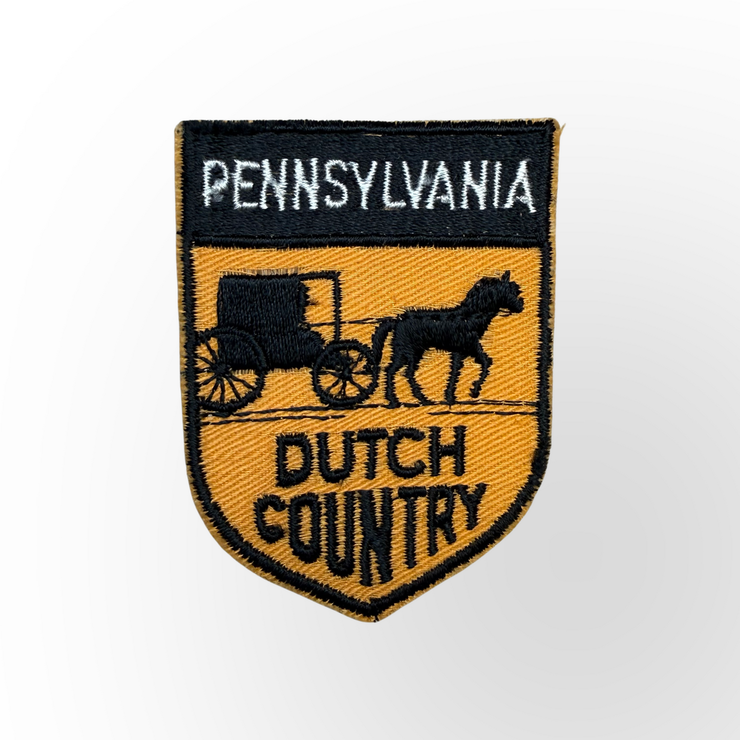 An embroidered shield-shaped patch featuring a black and gold design with the text 'PENNSYLVANIA' at the top and 'DUTCH COUNTRY' at the bottom, plus a black silhouette of a horse-drawn carriage and horse in the center.
