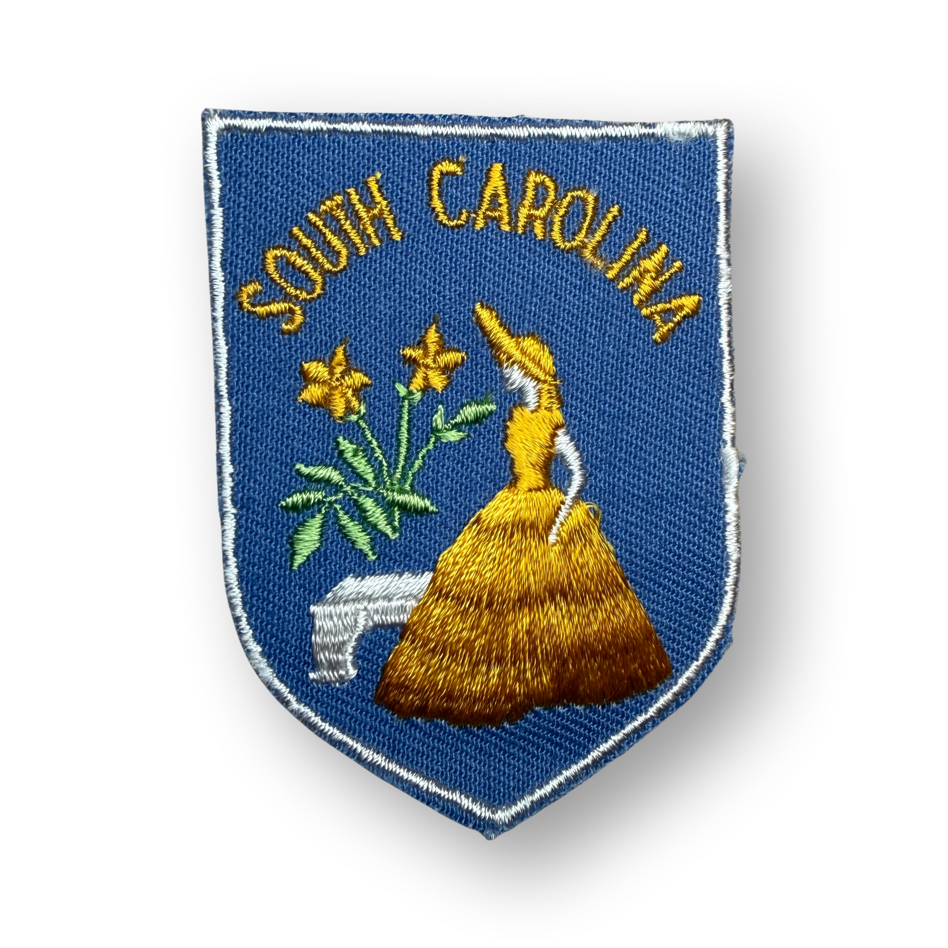An embroidered shield-shaped South Carolina state patch on a white background. The patch features a blue field with yellow embroidered text reading 'SOUTH CAROLINA' above a yellow bonnet, green leaves, and yellow flowers, bordered by a white stitched edge.