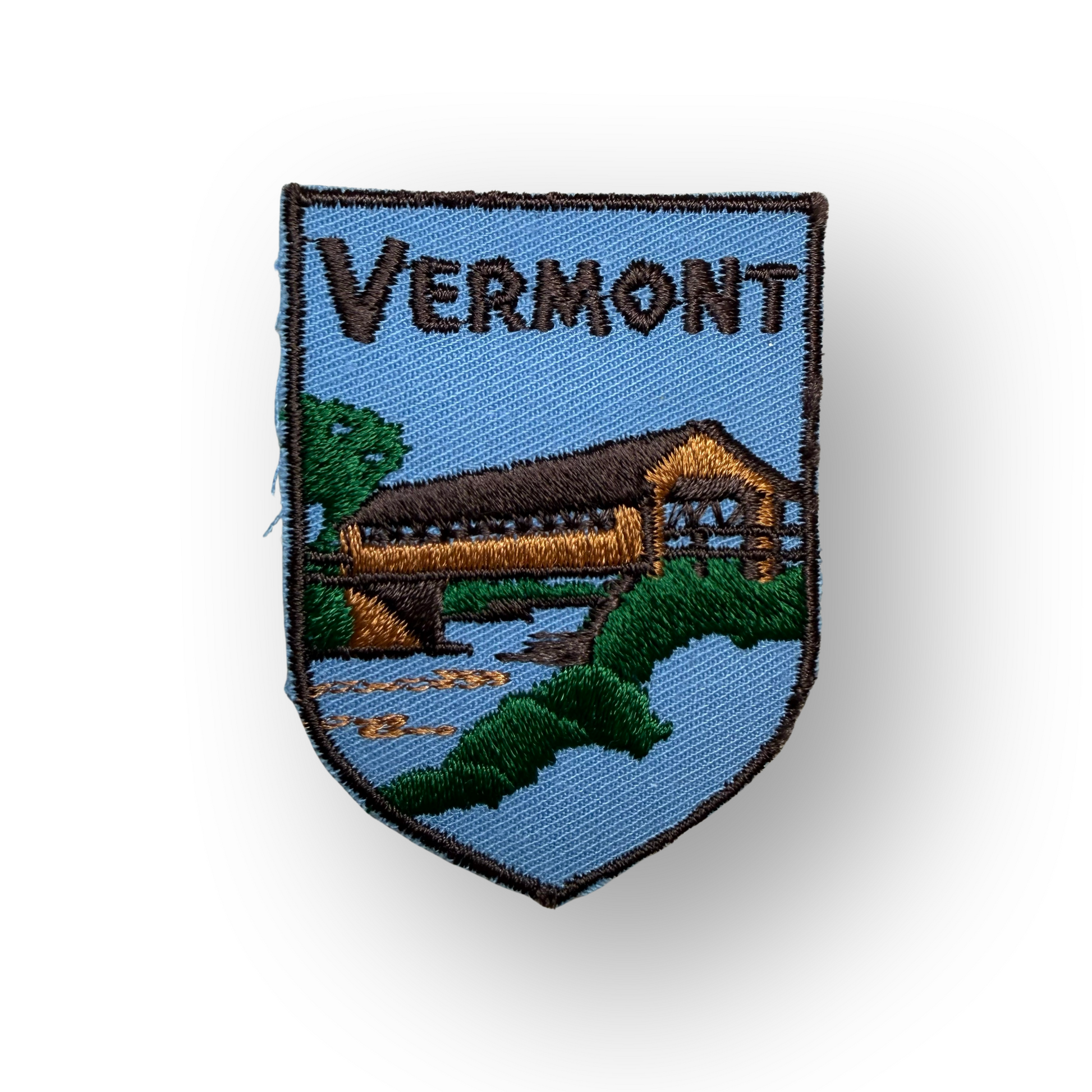 An embroidered shield-shaped patch featuring a covered bridge over water with green trees and the word 'VERMONT' at the top, shown on a white background with a soft shadow.