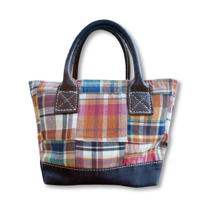 A small patchwork plaid tote bag with dark brown leather handles and a dark navy bottom base, shown on a plain white background.