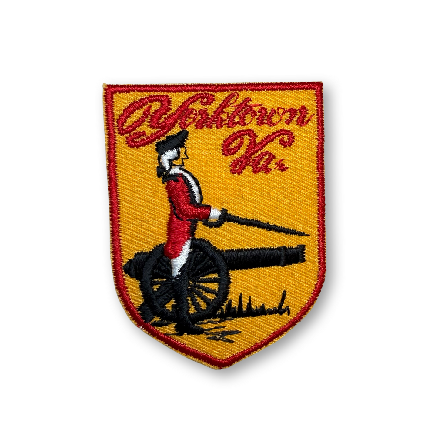 An embroidered shield-shaped patch featuring a red and white uniformed figure on a black cannon against a yellow background, with red script text at the top, shown on a plain white background.