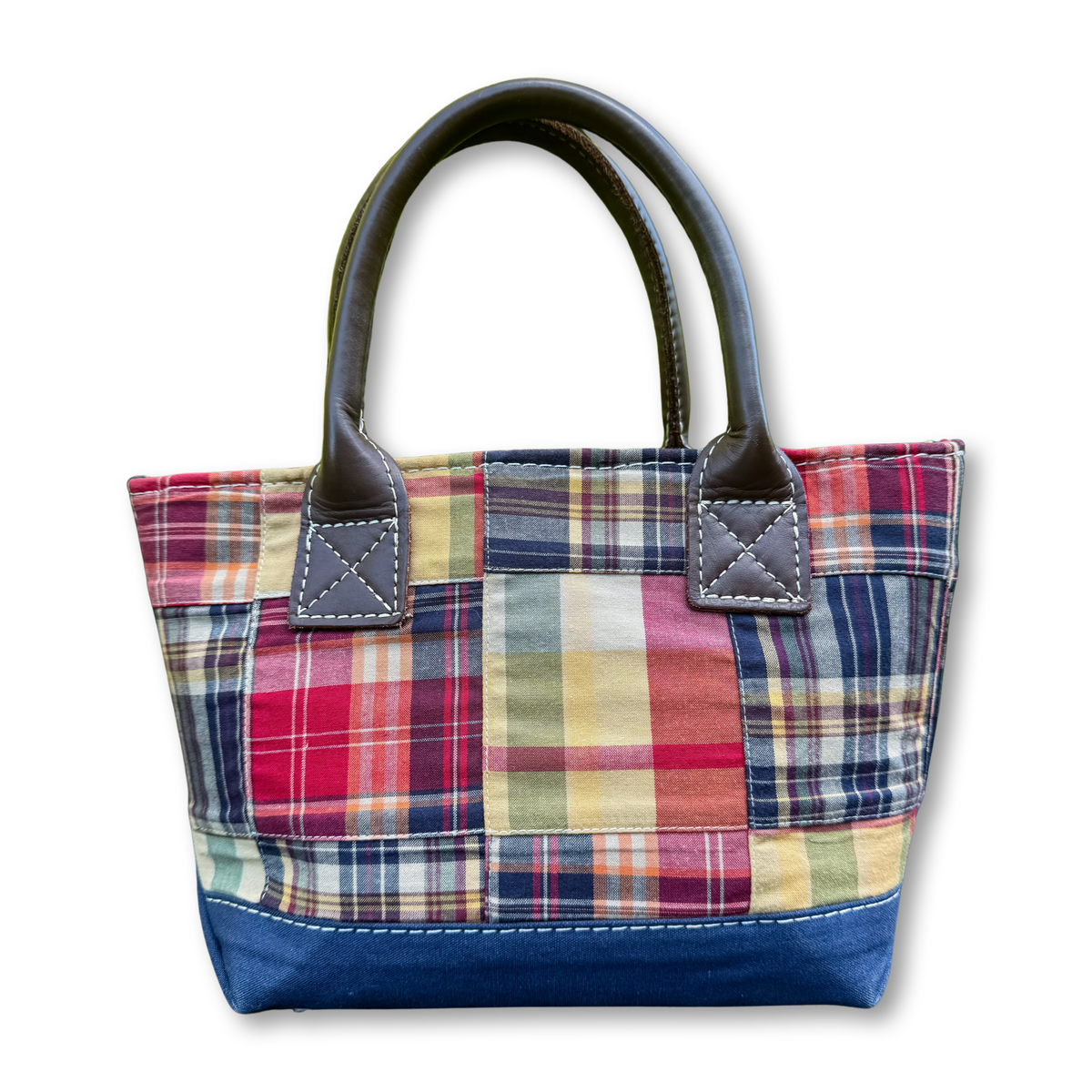 A patchwork plaid tote bag with dark brown leather handles and a solid navy blue bottom base, shown on a plain white background.