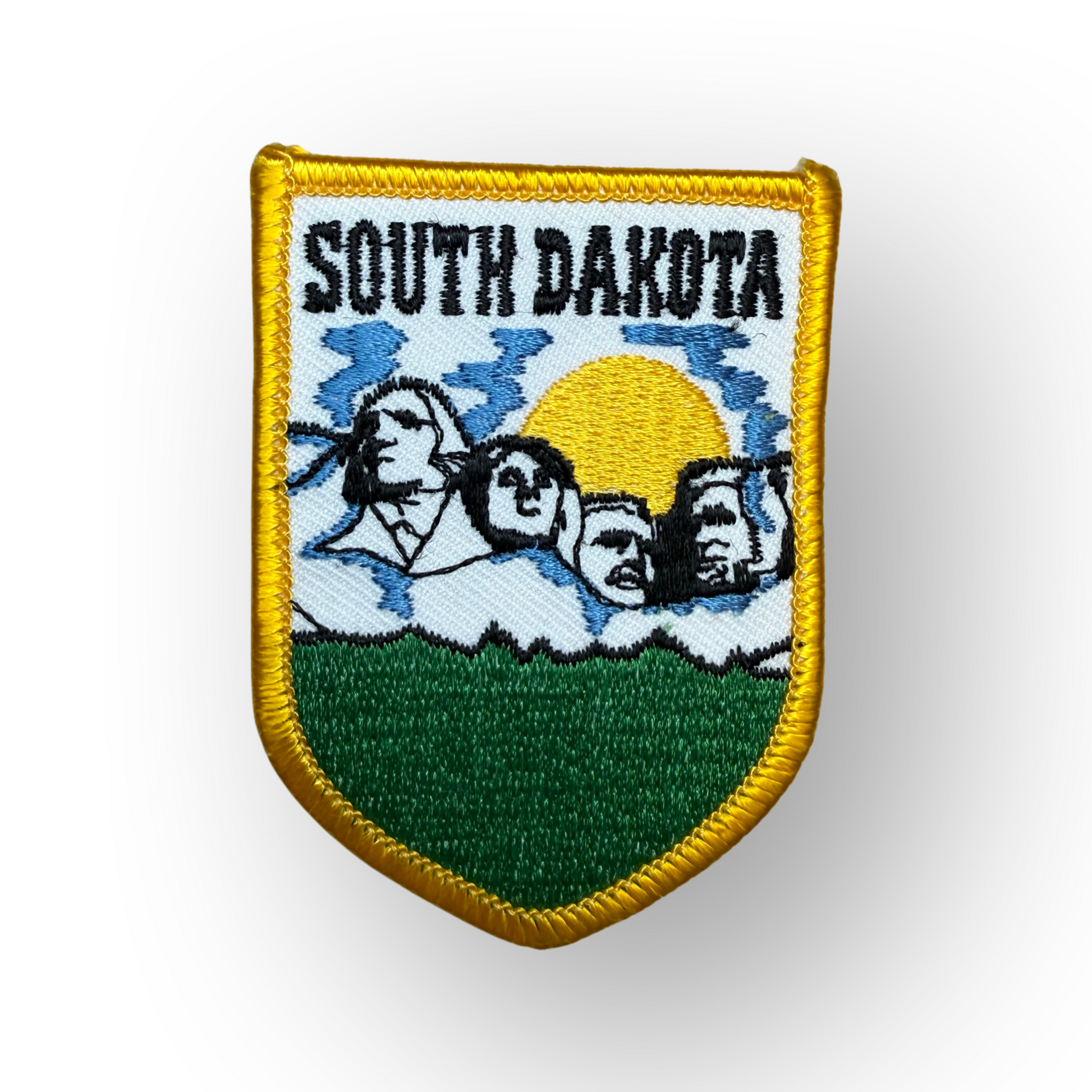 A shield-shaped embroidered patch featuring the text 'SOUTH DAKOTA' above a landscape design with blue sky, white clouds, a yellow sun, black mountain silhouettes, and a green field, all outlined with a gold border.