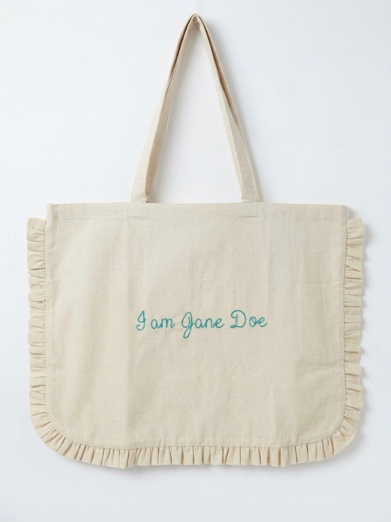 A cream-colored canvas tote bag with ruffled trim along the bottom and handles, featuring the text 'I am Jane Doe' embroidered in teal on the front, set against a plain white background.