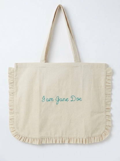 A cream-colored canvas tote bag with ruffled trim along the bottom and handles, featuring the text 'I am Jane Doe' embroidered in teal on the front, set against a plain white background.