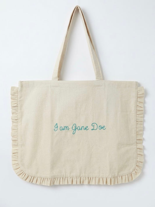 A cream-colored canvas tote bag with ruffled trim along the bottom and handles, featuring the text 'I am Jane Doe' embroidered in teal on the front, set against a plain white background.