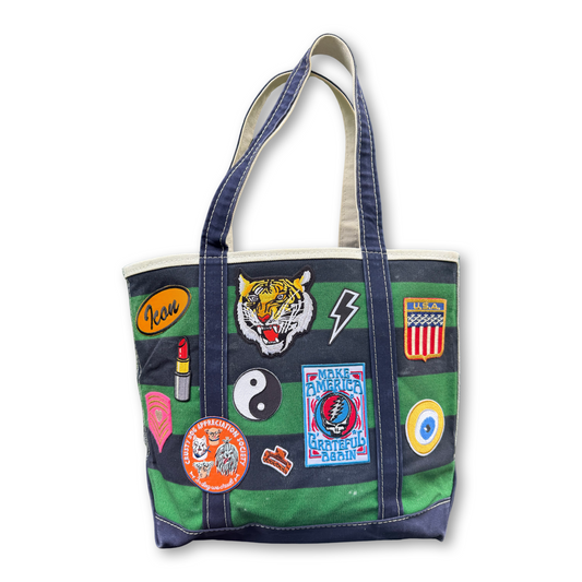 A canvas tote bag with a green and navy striped pattern, featuring multiple colorful patches on the front, including a tiger patch, a lightning bolt patch, a yin-yang patch, and a Grateful Dead patch, set against a plain white background.