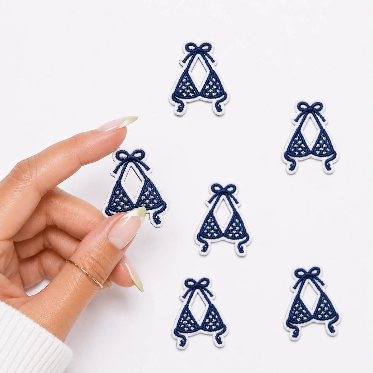 A hand with manicured nails holds one embroidered patch while five identical navy blue bikini patch designs are arranged on a clean white background.