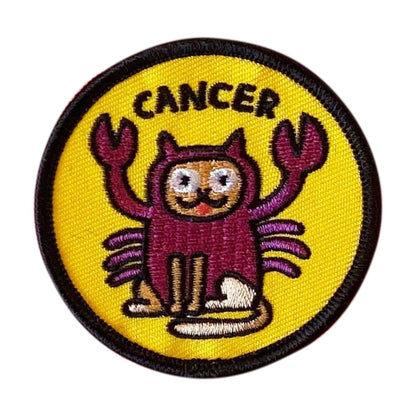 A circular embroidered patch featuring a cartoon crab character with the word 'CANCER' above it, on a bright yellow background with a black stitched border.