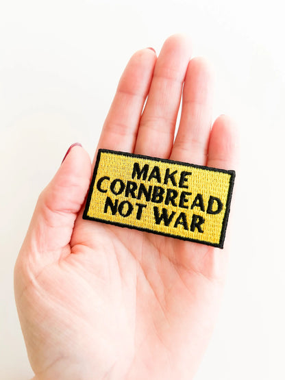 Hand holding a patch with 'MAKE CORNBREAD NOT WAR' text on a white background