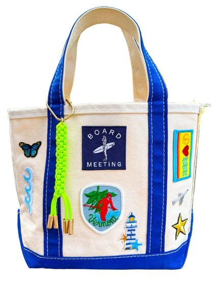 Beige tote bag with colorful patches and a blue handle, featuring a 'Board Meeting' patch.