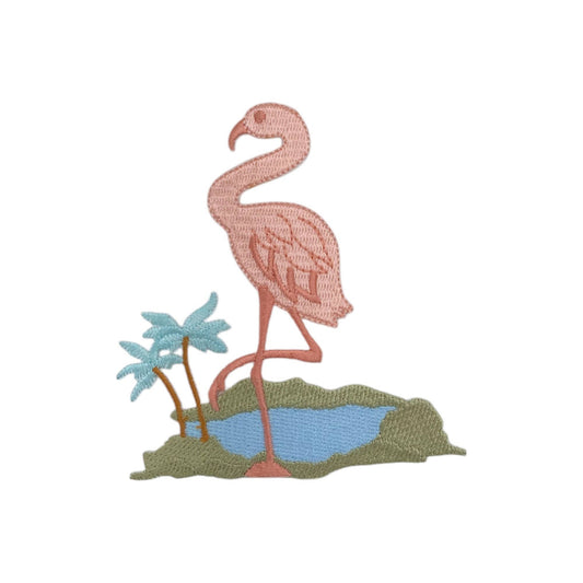 An embroidered patch featuring a pink flamingo standing on a patch of green grass next to a small blue pond, with two light blue palm trees, on a plain white background.