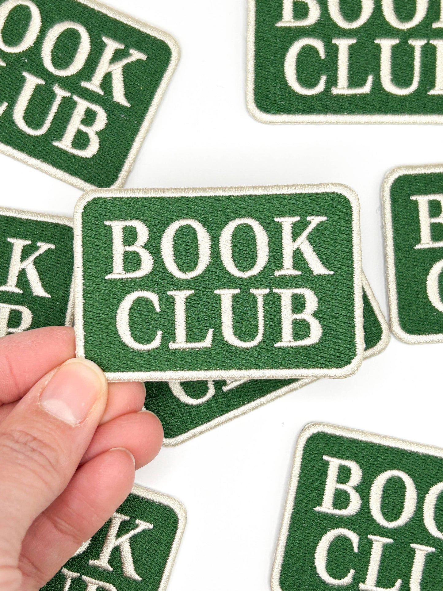 Green patches with 'BOOK CLUB' text held by a hand on a white background