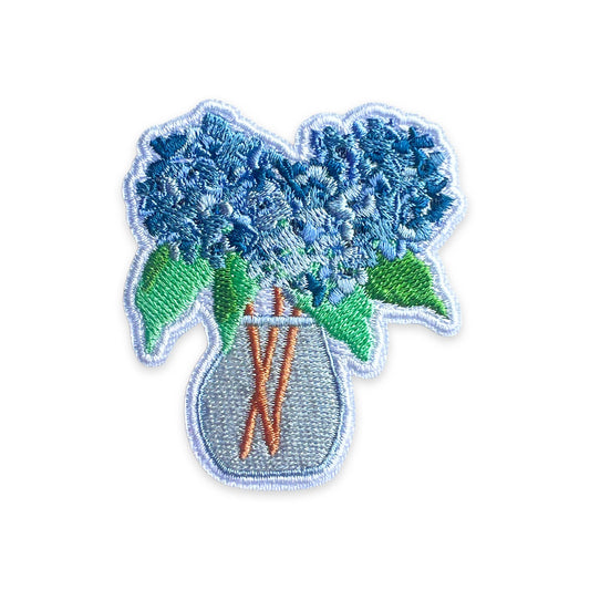An embroidered patch depicting blue hydrangea flowers with green leaves in a light blue vase, set against a plain white background.