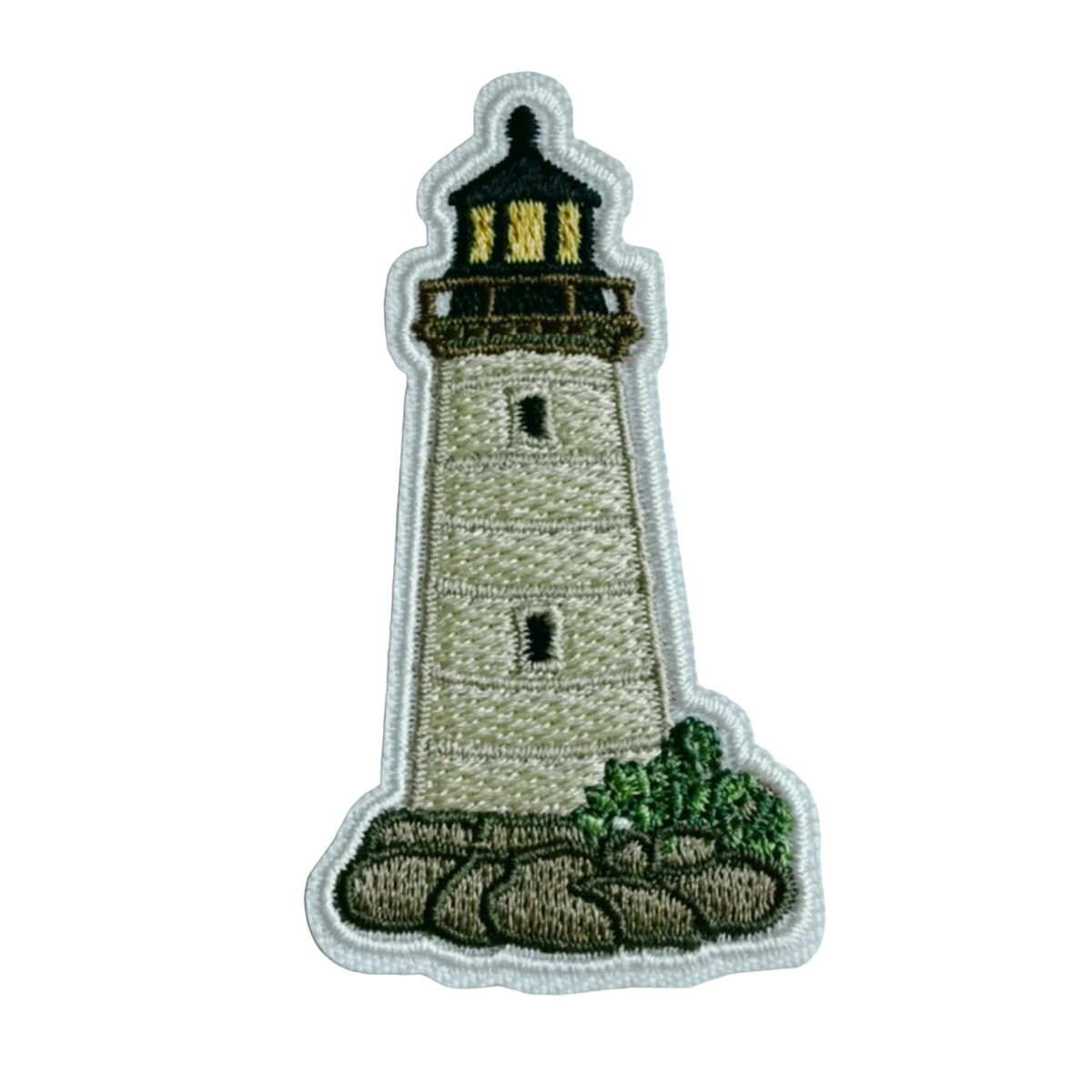 An embroidered patch of a lighthouse on a rock outcrop, featuring a beige tower, black and yellow lighthouse top, green foliage, and brown rocks, all outlined in white stitching, shown on a light gray textured background.