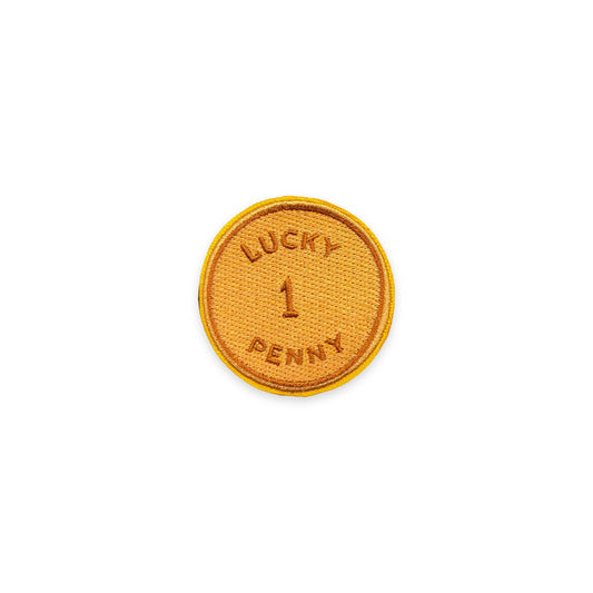 A circular embroidered patch with a golden-yellow border and a textured orange-brown center, featuring the text 'LUCKY 1 PENNY' embroidered in matching thread on a plain white background.