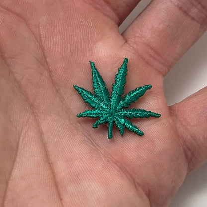 A metallic green embroidered cannabis leaf patch resting on a person's palm to show its scale.